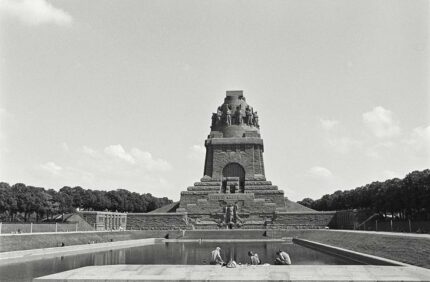 Das Völkerschlachtdenkmal in Leipzig mit Wasserbecken. Davor sitzen einige junge Leute.